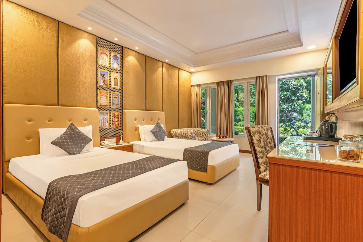A hotel room with two beds, a sitting area, a desk with a coffee maker, and a view of greenery outside.