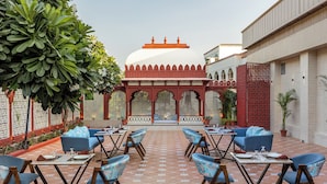 A courtyard with blue seating and tables, a white building with arches, and a red lattice structure. - Sterling Amargarh Jodhpur (Luni)