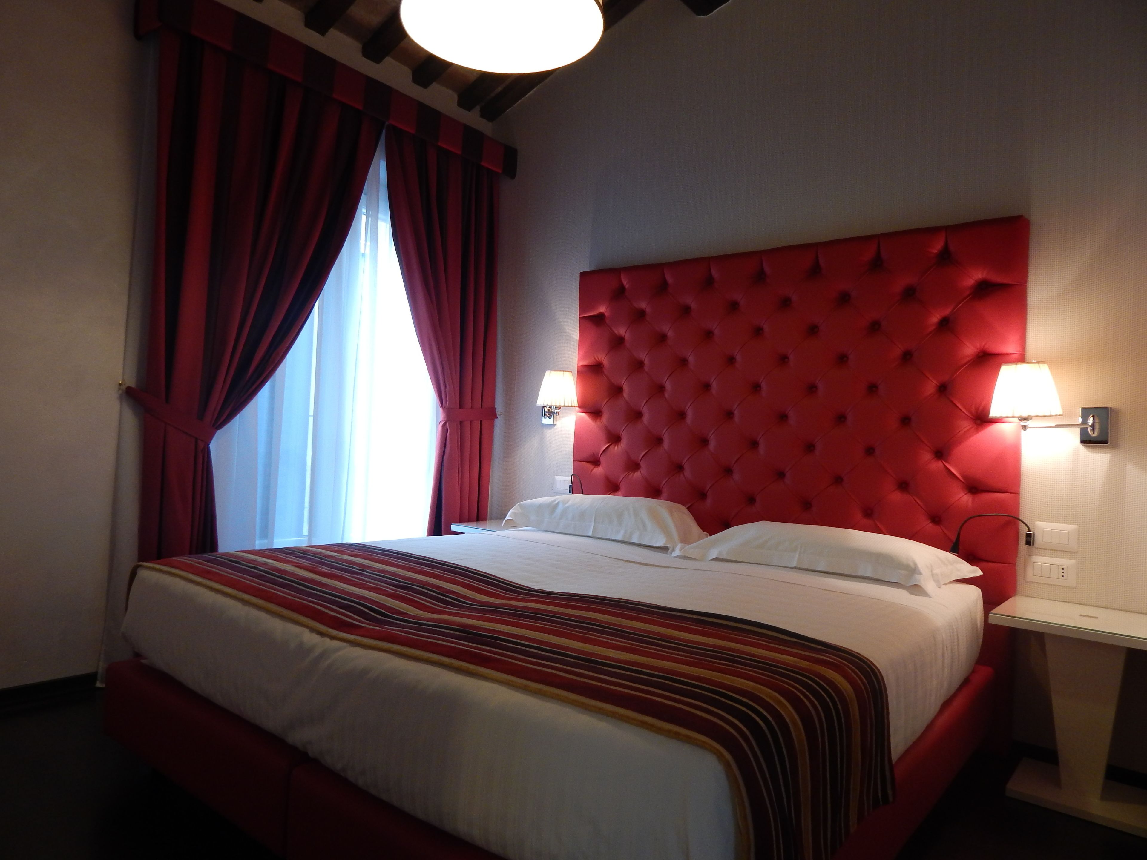 Una habitación de hotel con una cama grande, cortinas rojas, un cabecero rojo y dos lámparas de noche.