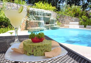 Een bord met guacamole, tortilla chips en een partje limoen, met een zwembad en waterval op de achtergrond.