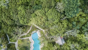 Luchtbeeld van een resort met een zwembad, omgeven door weelderige begroeiing, een verhard parkeerterrein en een grindpad.