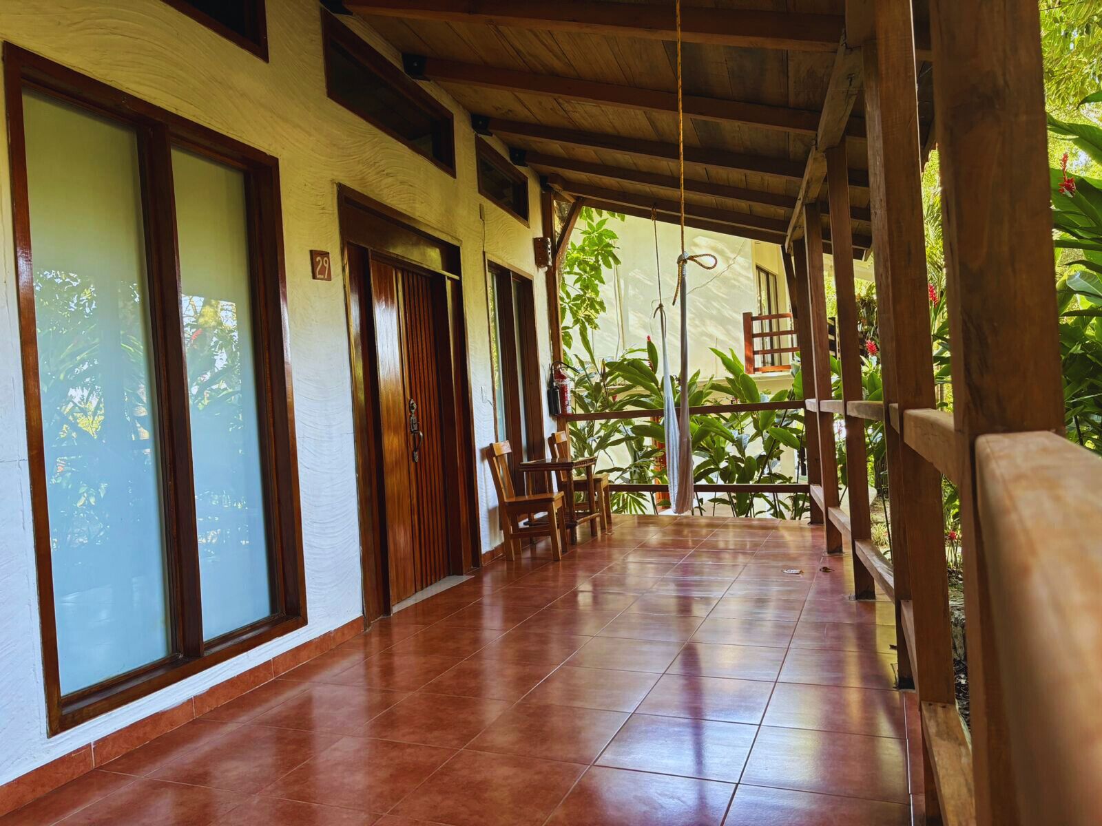 Um corredor coberto com piso de azulejos, portas de madeira e vista para vegetação exuberante.
