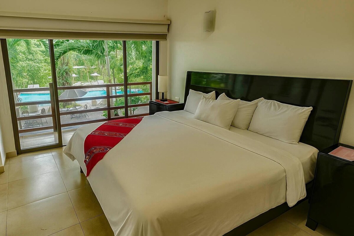Une chambre d’hôtel avec un grand lit, une table de chevet, une fenêtre donnant sur une piscine et un espace verdoyant, et un ventilateur de plafond.