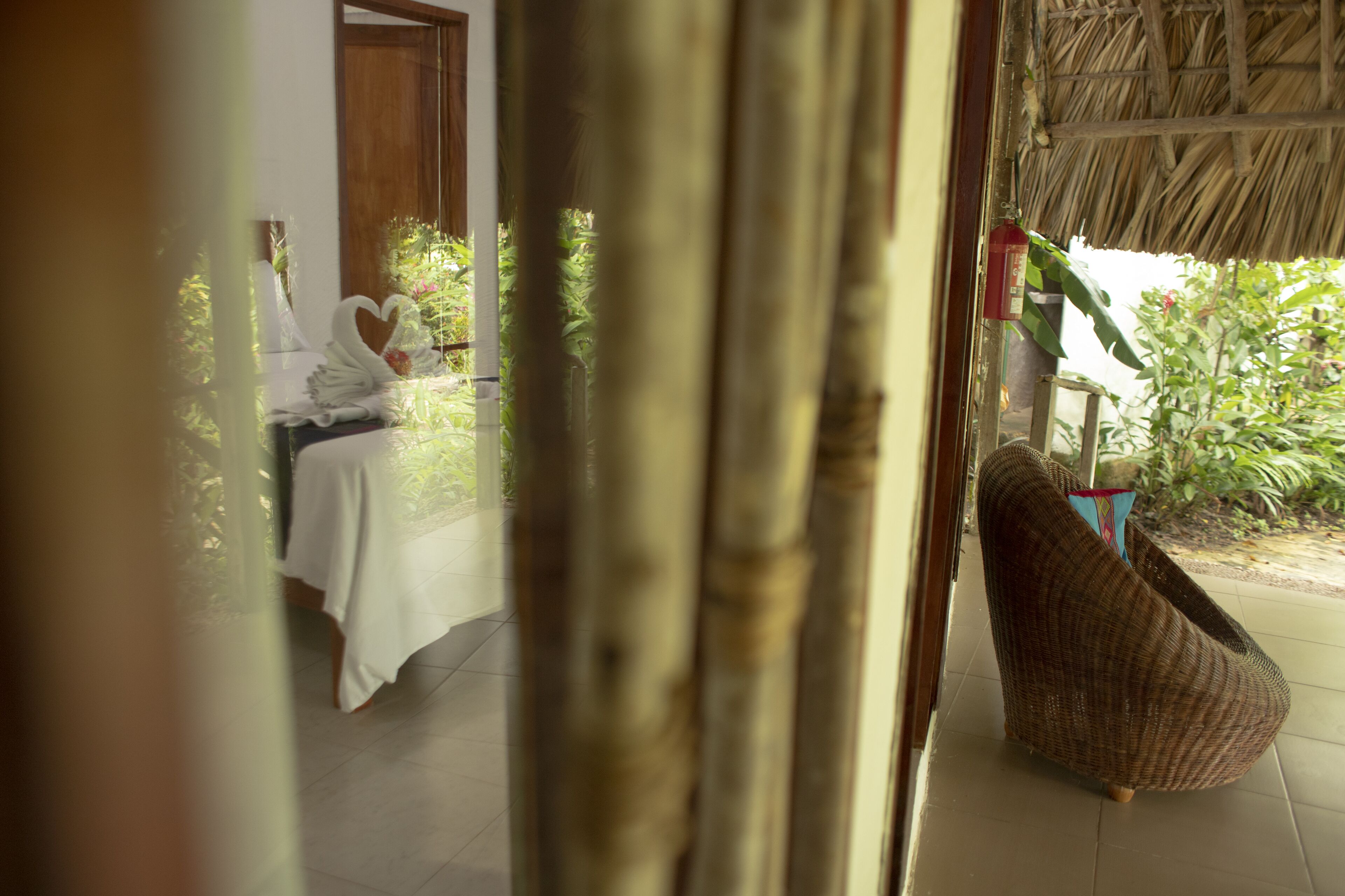 A room with a bed, a wicker chair, and a thatched roof outside.