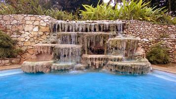 Una piscina de piedra con cascada y agua en movimiento, rodeada de una pared de piedra y abundante vegetación.