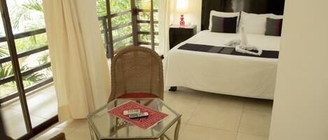 A hotel room with a bed, a glass table with a remote control, a wicker chair, and a view of greenery through the window.
