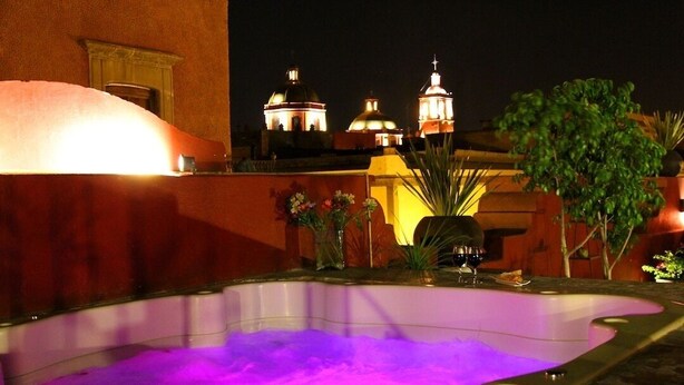 Un bain à remous éclairé d’une lumière violette, entouré de plantes en pot et offrant une vue sur les bâtiments de la ville la nuit.