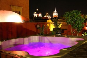 Un bain à remous éclairé d’une lumière violette, entouré de plantes en pot et offrant une vue sur les bâtiments de la ville la nuit.