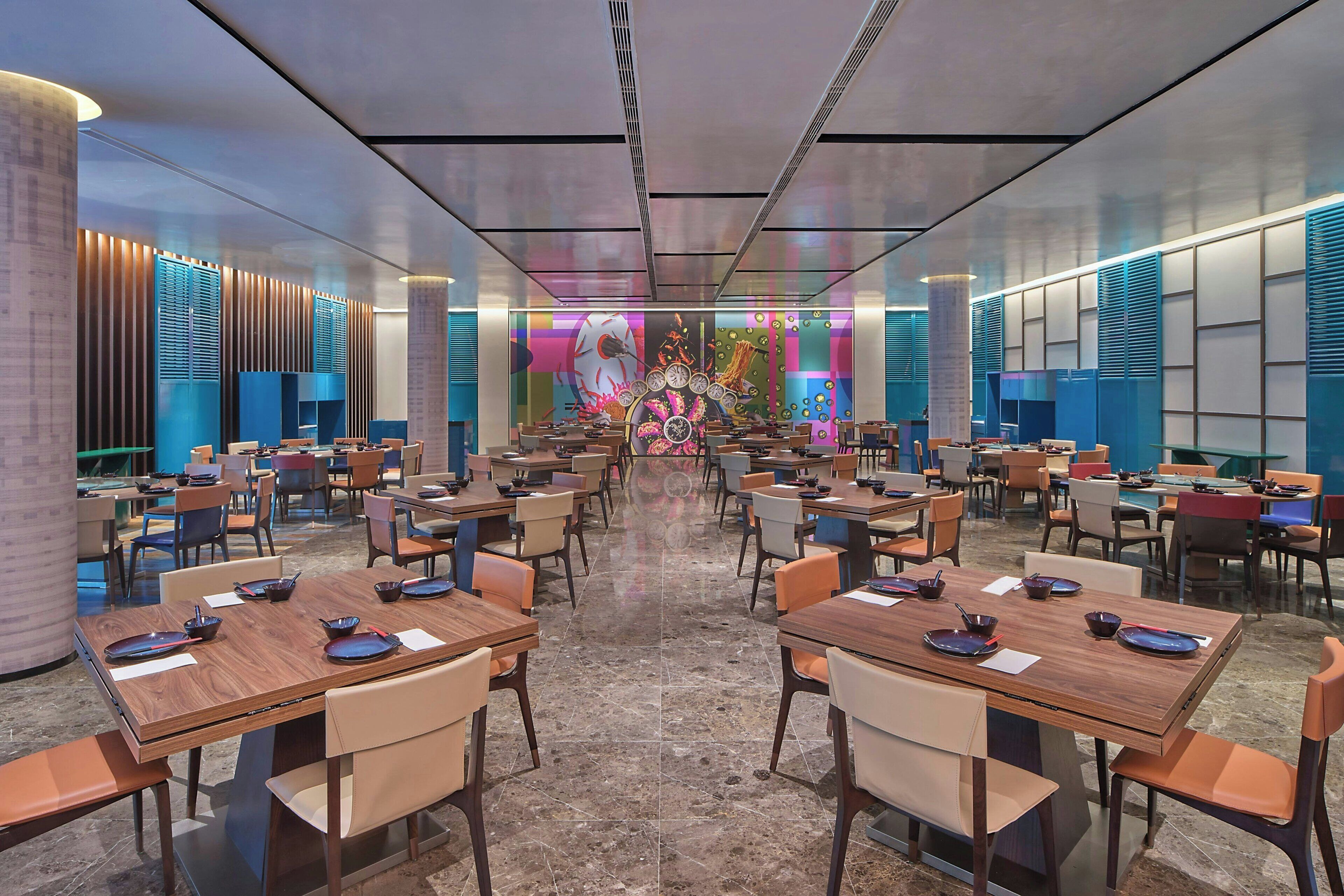 Un amplio vestíbulo de hotel con varias mesas y sillas de comedor, una pared decorativa central y un diseño de techo moderno.