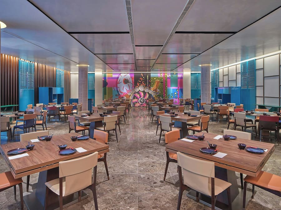 Un amplio vestíbulo de hotel con varias mesas y sillas de comedor, una pared decorativa central y un diseño de techo moderno.