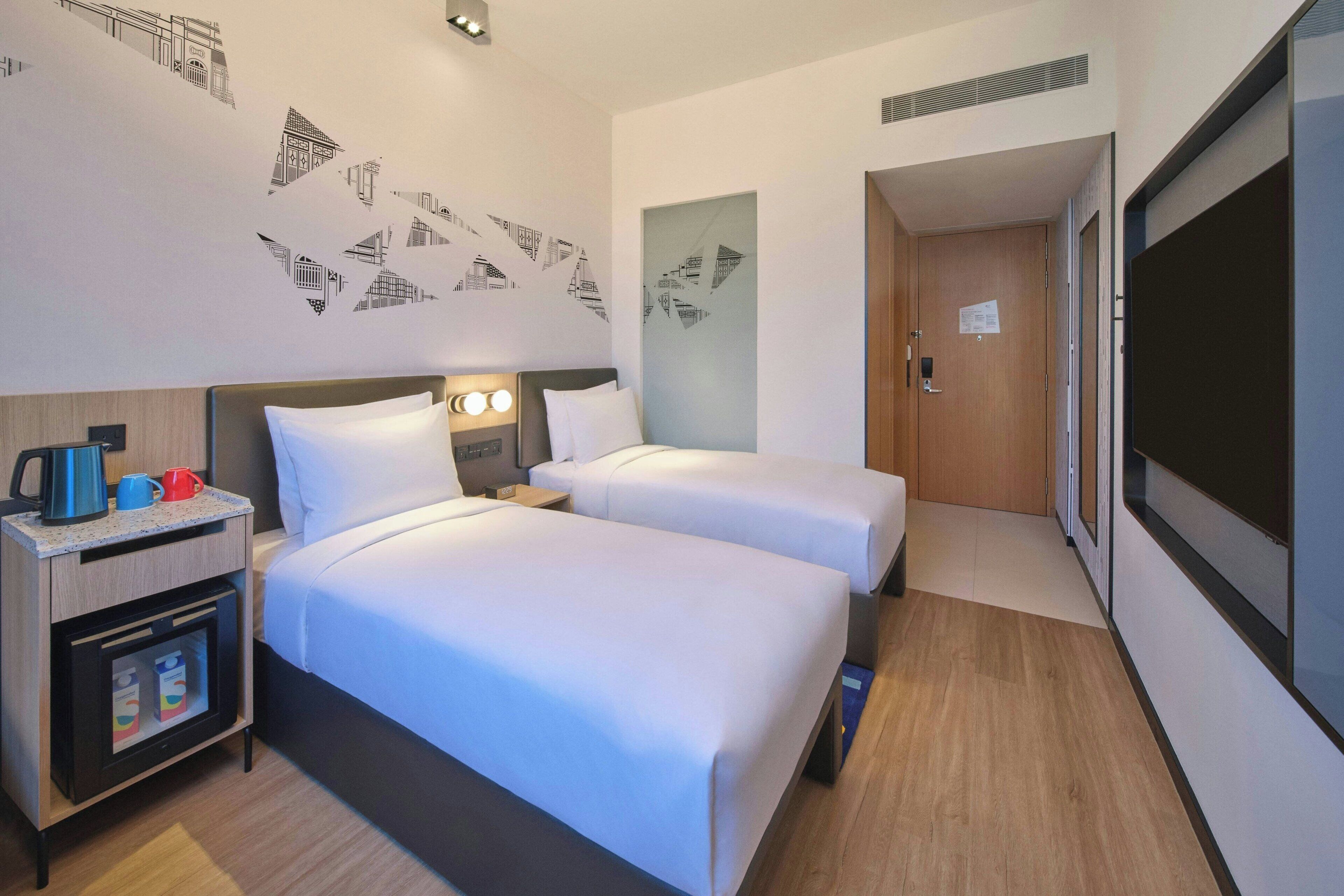 Une chambre d’hôtel avec deux lits, une table de chevet avec une machine à café, un mini-réfrigérateur et un mur orné de motifs géométriques.