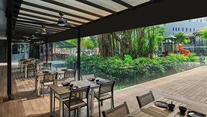 Un área de comedor al aire libre con mesas y sillas, barandilla de vidrio y vistas a un parque infantil y zonas verdes.