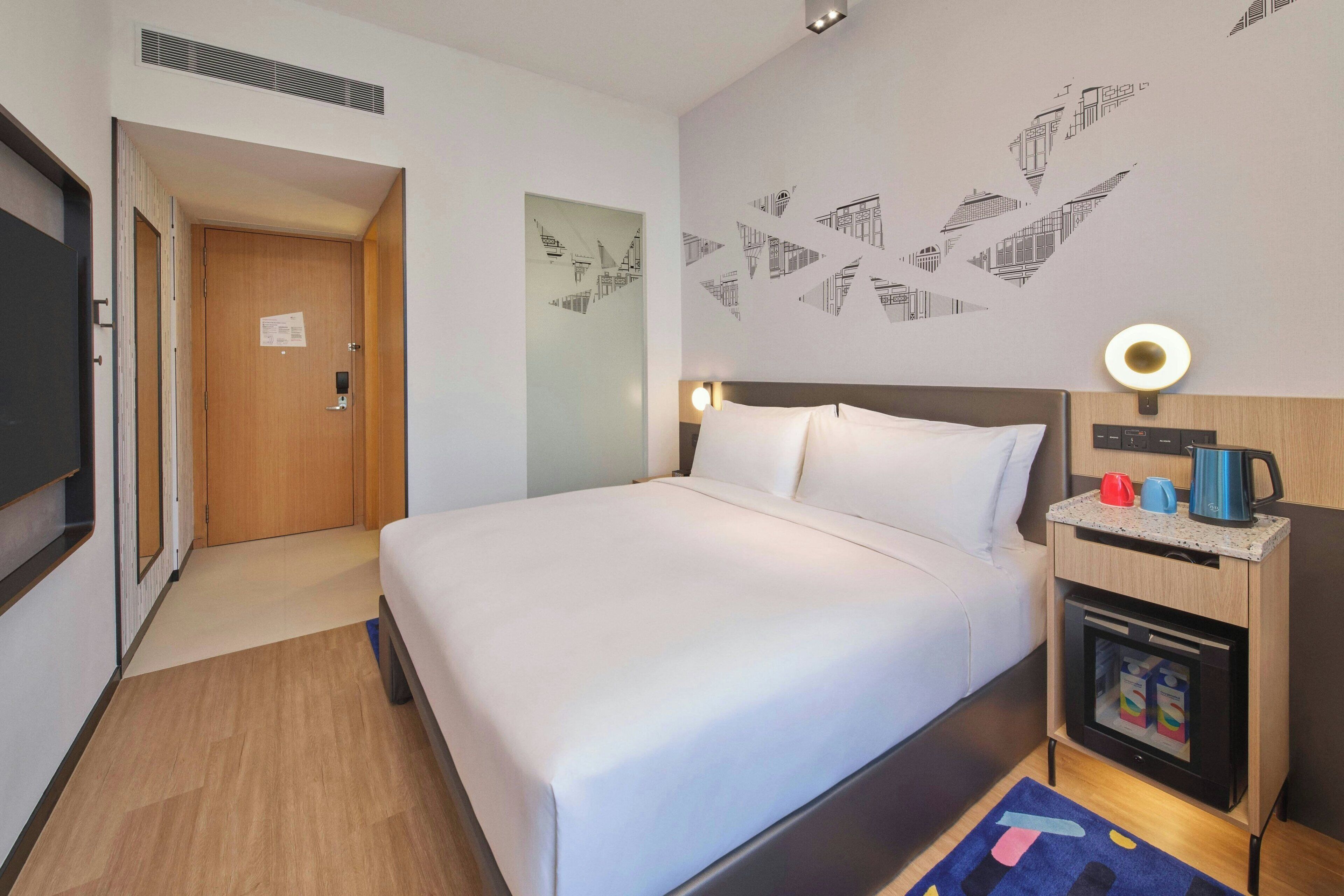 Hotelkamer met een bed, een nachtkastje met een lamp, een kastje met een koffiezetapparaat en een muur met geometrische patronen.