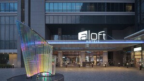 Una entrada de hotel moderna con un llamativo letrero de 'Alloft Hotels', una gran escultura iluminada y un área de recepción amplia.