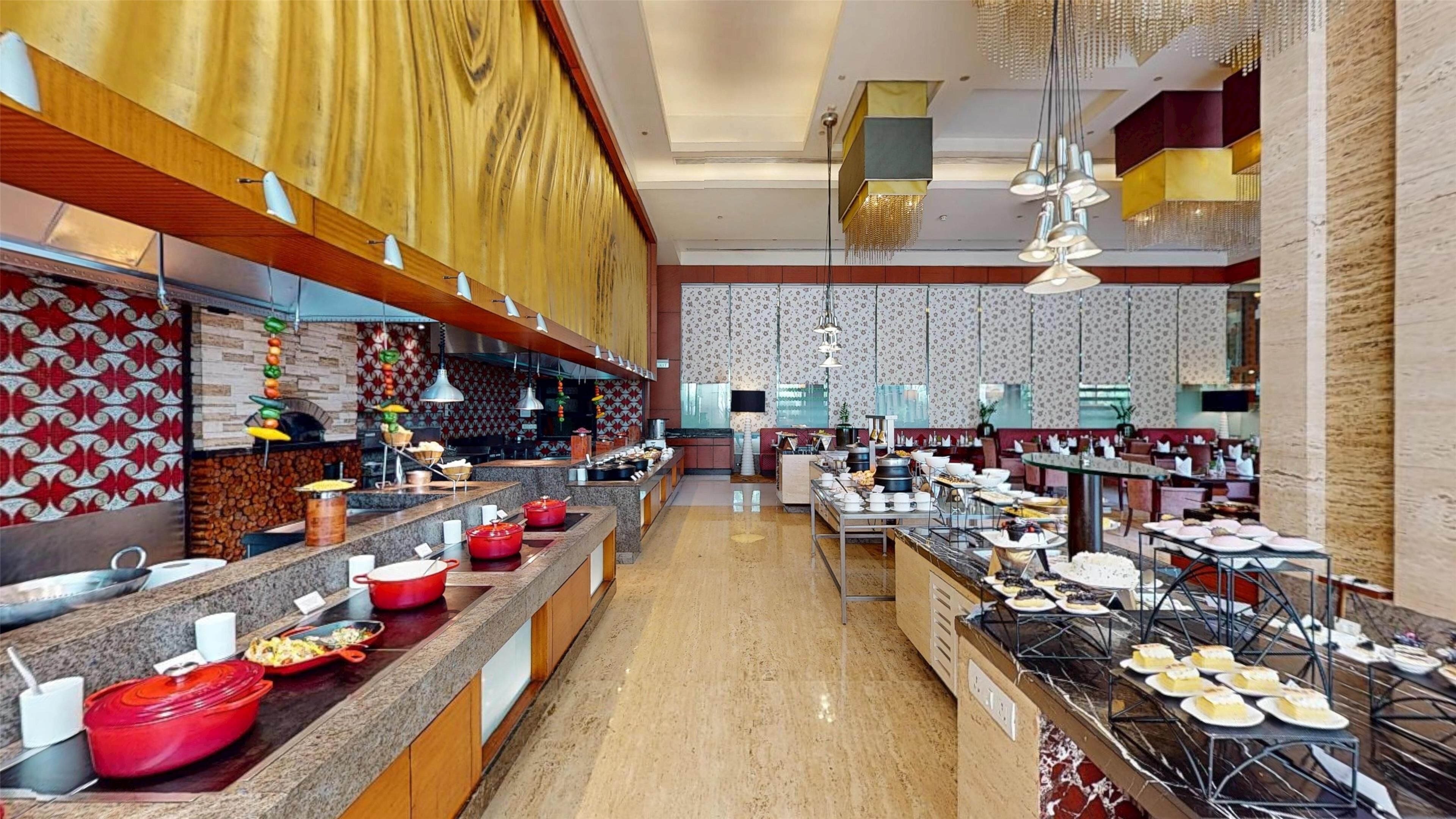 A spacious hotel dining area with multiple buffet stations, a central serving area, and a variety of seating arrangements.