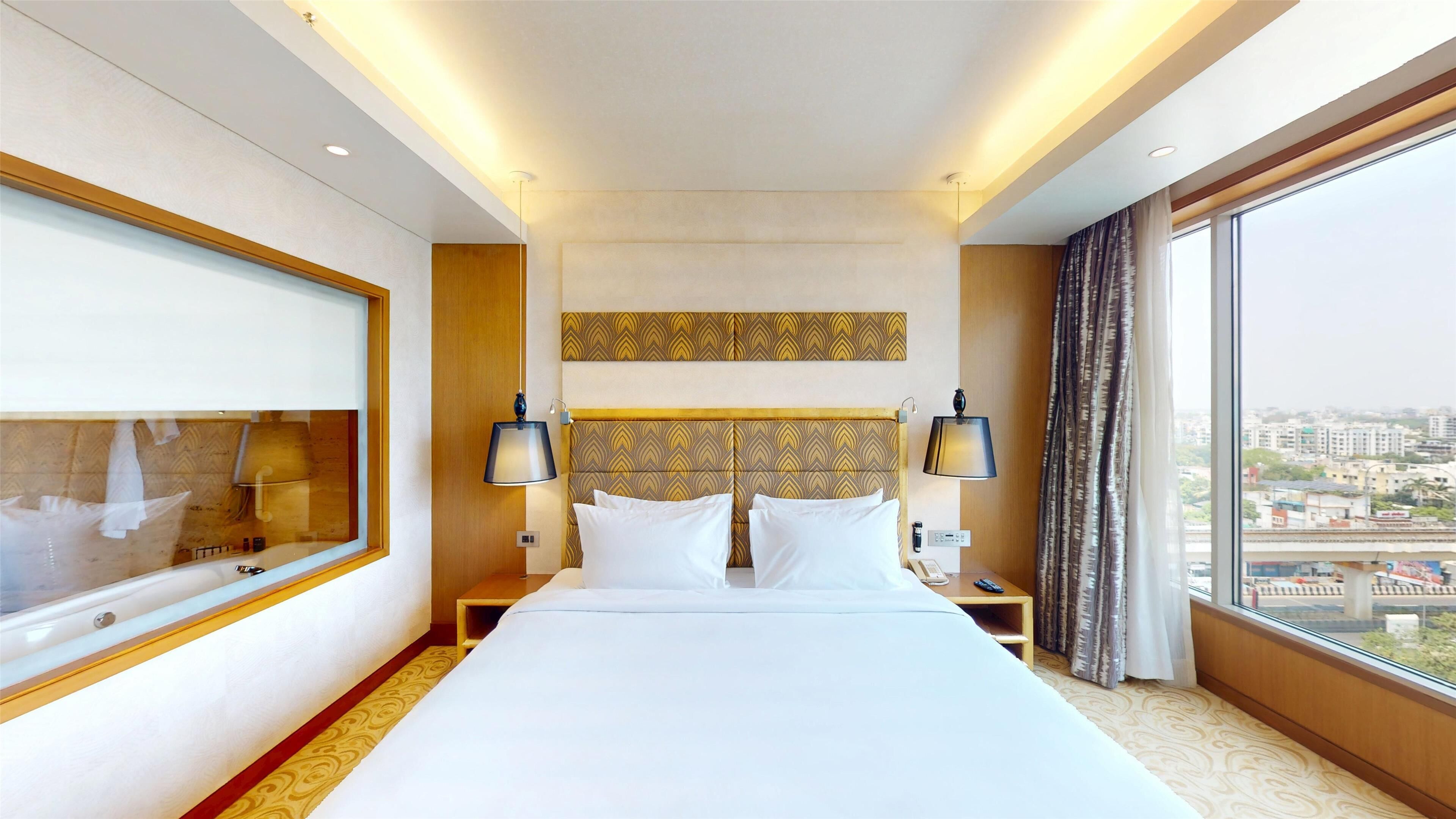A hotel room with a large bed, two bedside tables, a nightstand, a mirror, and a large window with a city view.
