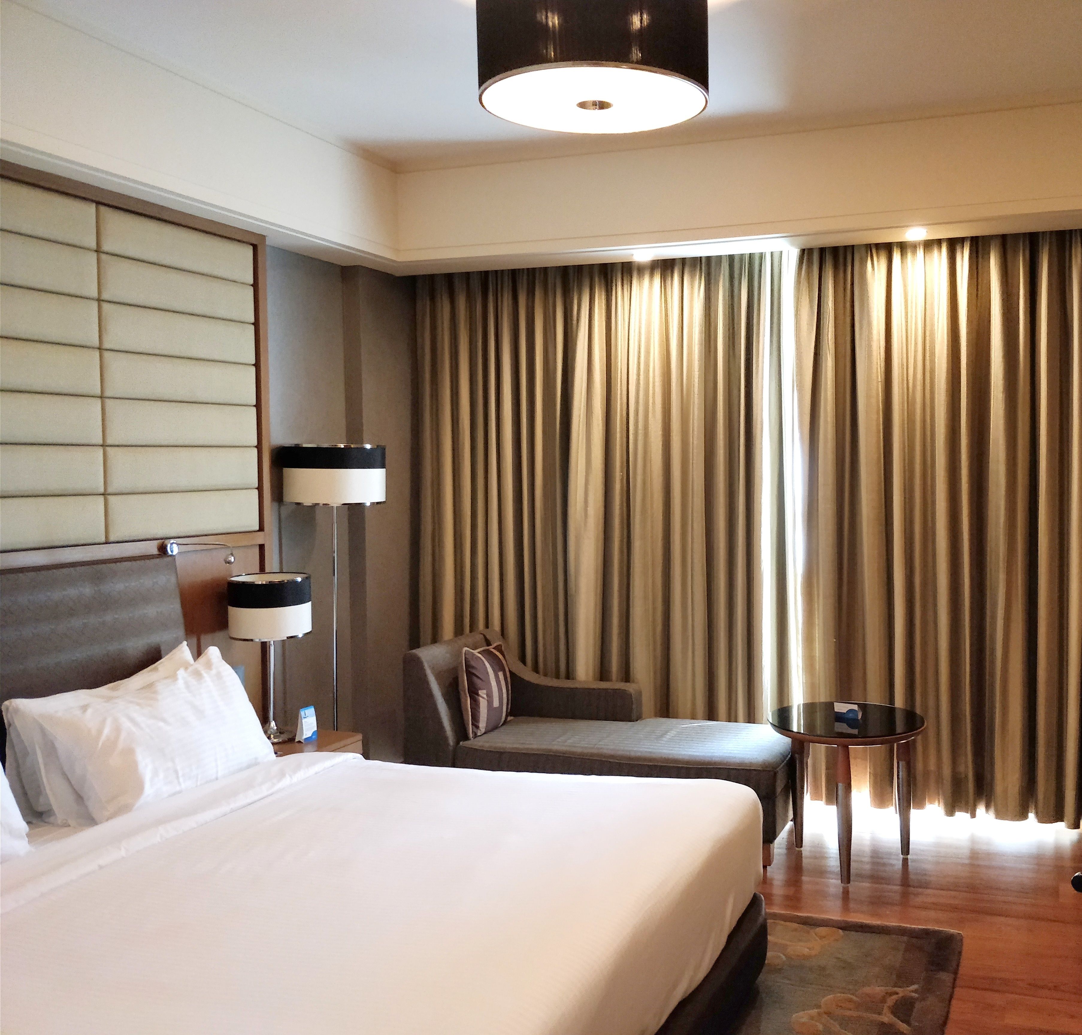 Une chambre d’hôtel avec un grand lit, une table de chevet avec une lampe, une petite table ronde, un canapé et une vue sur les rideaux.