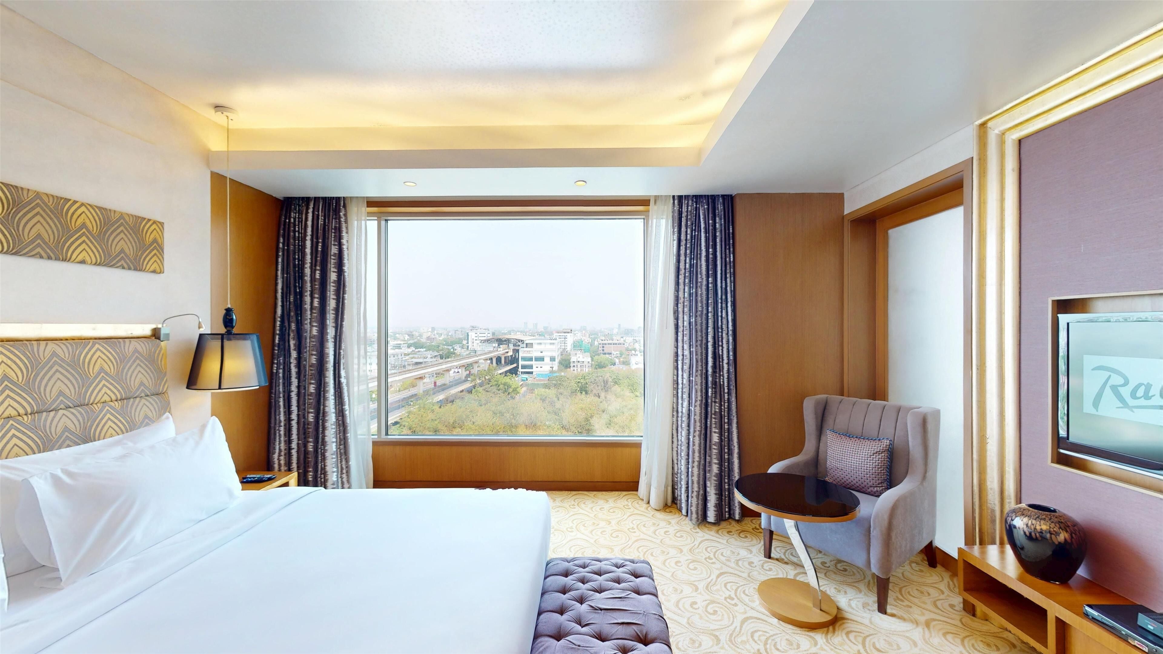 Habitación de hotel con una cama grande, un área de descanso con una silla y una mesita, televisión y vistas a la ciudad desde la ventana.