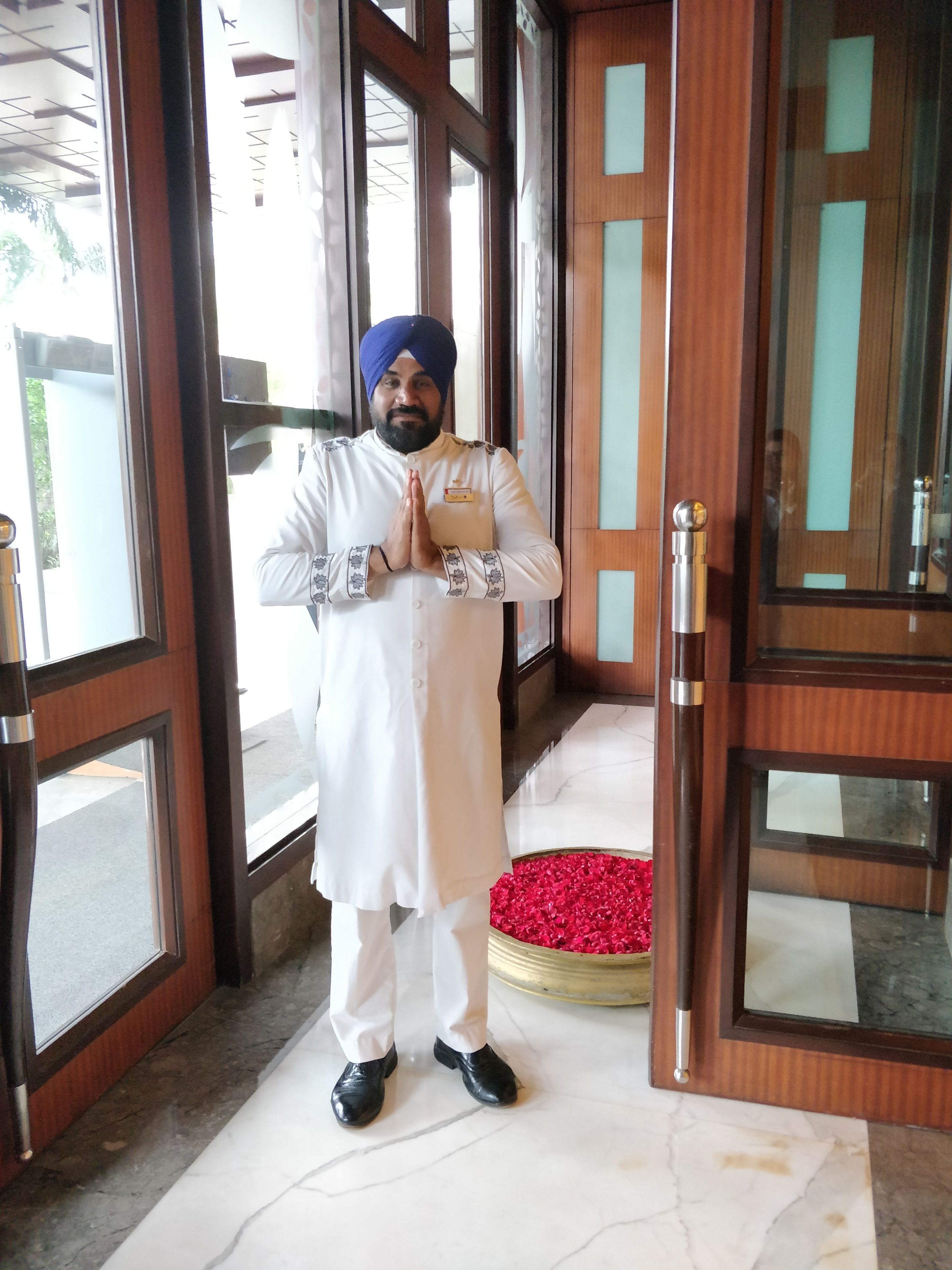 A person in traditional attire standing at a hotel entrance with a name tag, a prayer gesture, and a red carpet.