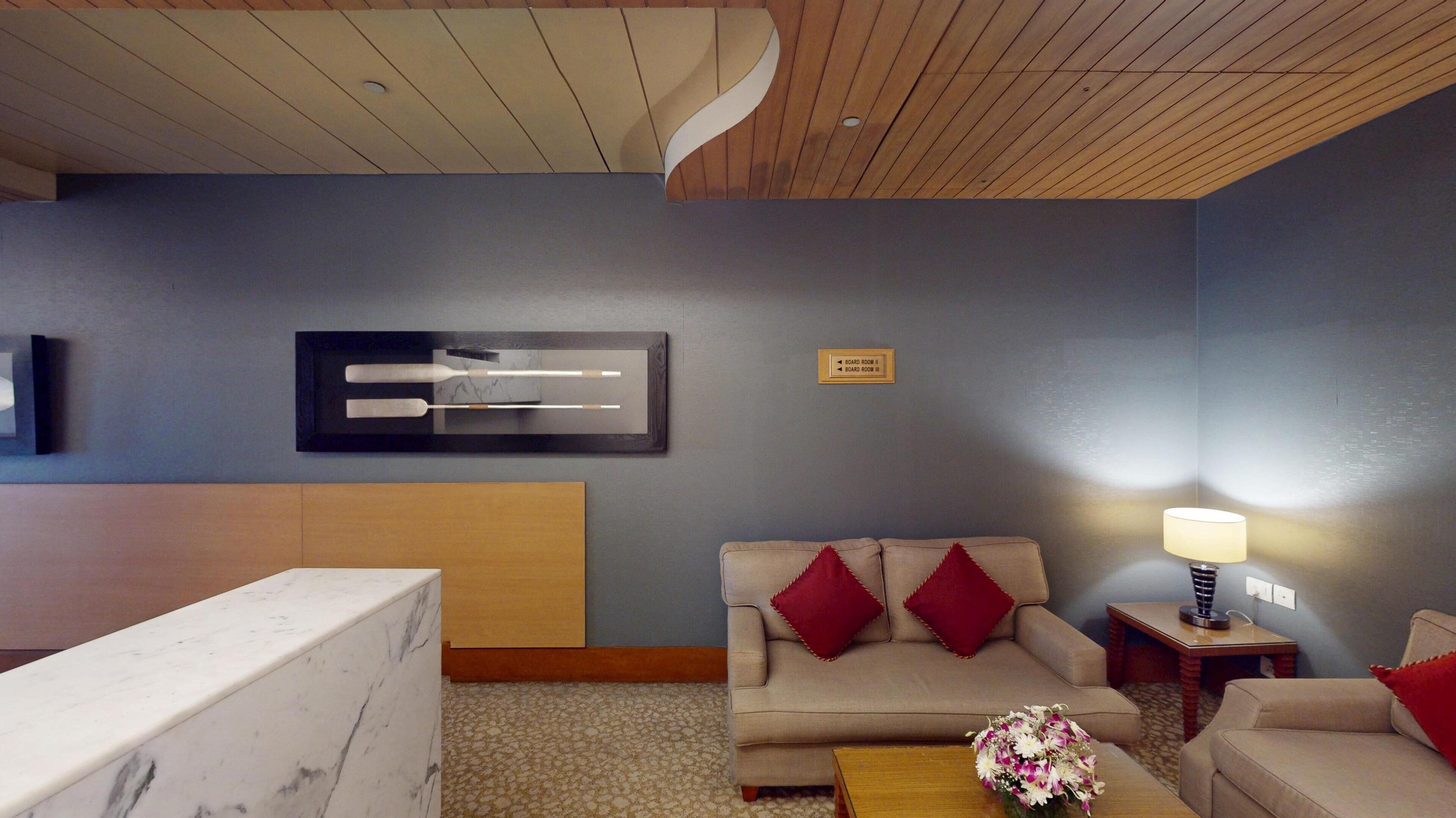 Una habitación de hotel moderna con un mostrador de recepción de mármol, un sofá beige con cojines rojos, una mesa de centro de madera y una obra de arte colgada en la pared.