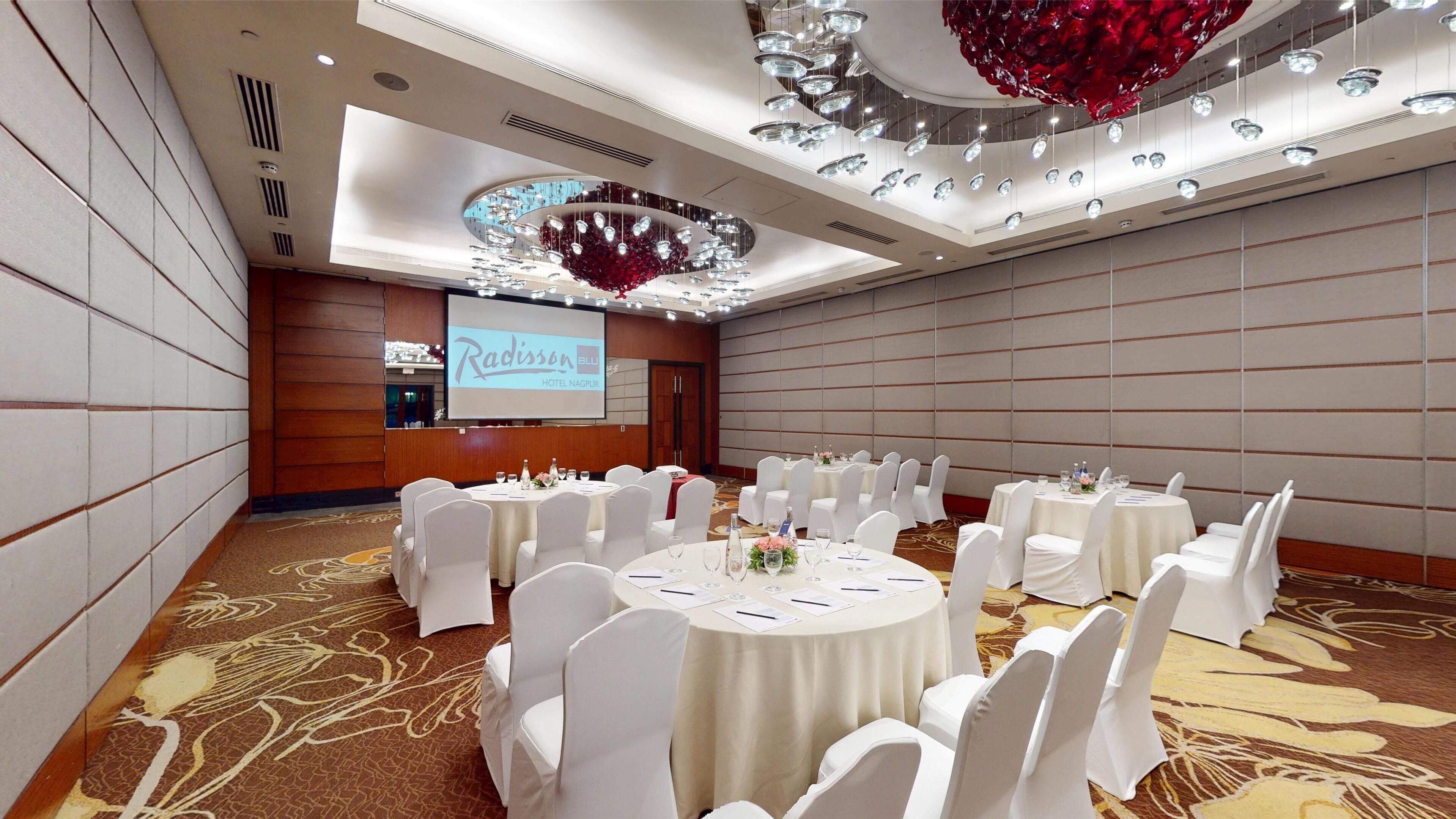 A conference room with round tables, white chairs, and a large screen displaying 'Radisson Hotel Nagpur'.