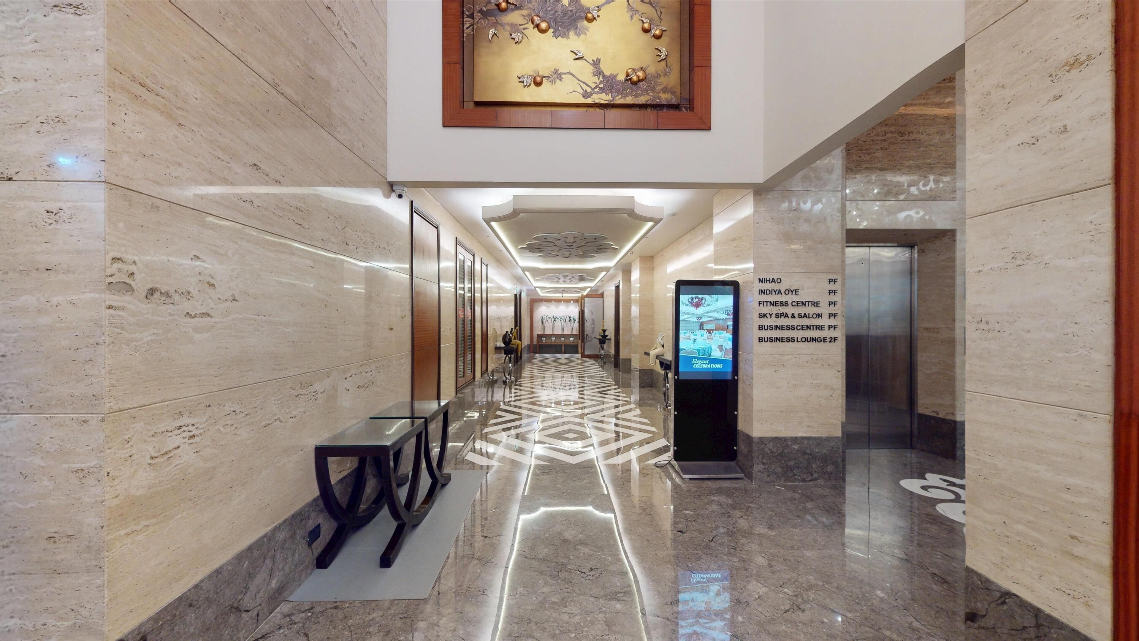 A hotel corridor with a polished marble floor, a reception desk, a digital information board, and a wall-mounted sign listing hotel amenities.