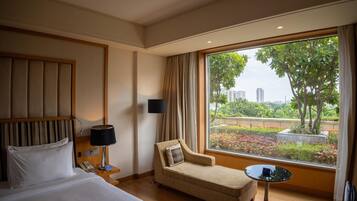 Quarto de hotel com cama, sofá, mesa pequena, luminária e grande janela com vista para áreas verdes e edifícios.