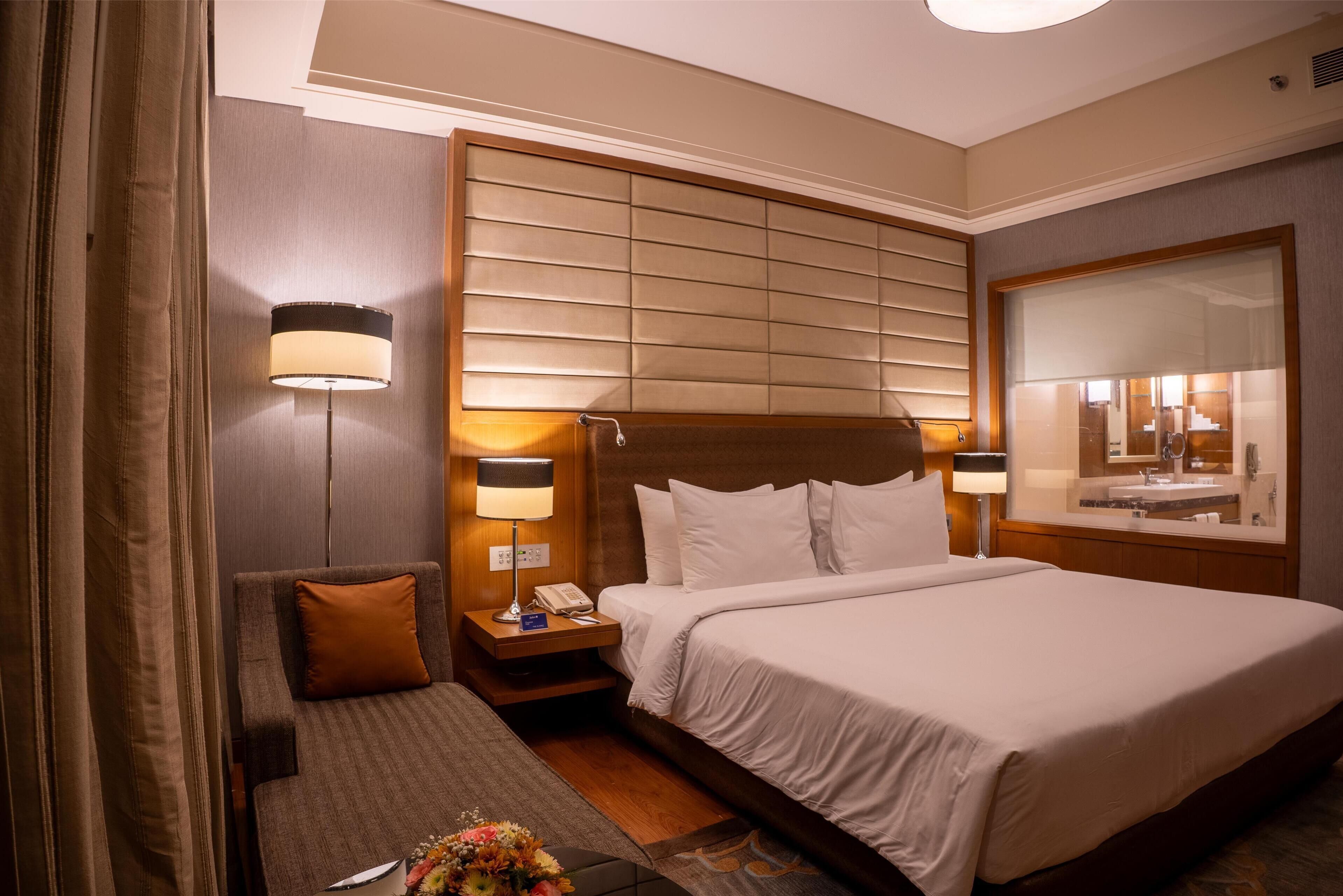 Camera d'albergo con un letto grande, un comodino con una lampada, un'area salotto con un cuscino e uno specchio sopra il letto.