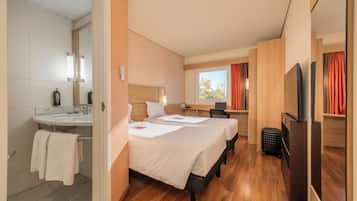 A hotel room with a bathroom, two beds, a desk, a TV, and a window with curtains.
