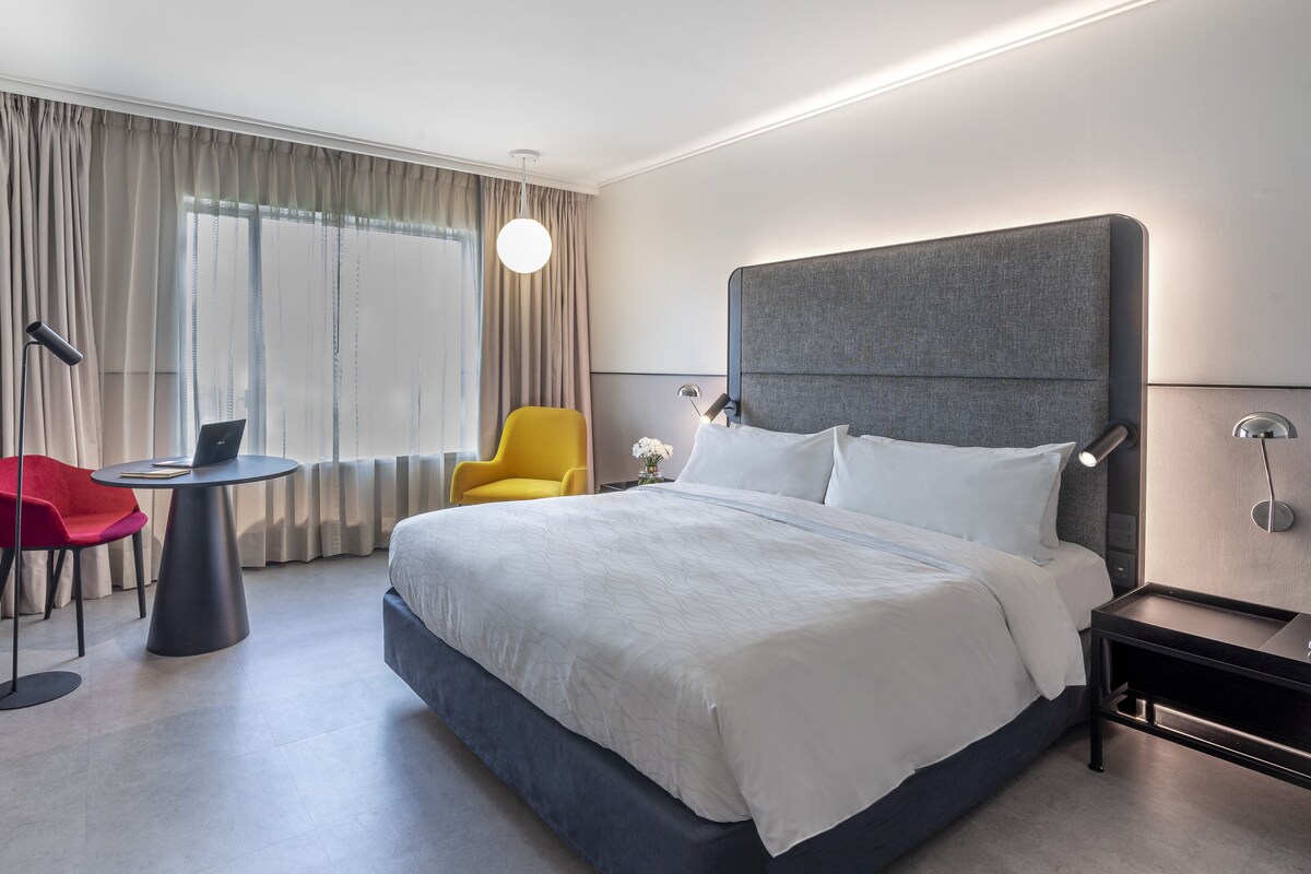 Una habitación de hotel moderna con una cama grande, un escritorio con una computadora portátil, una silla roja y un sillón amarillo.