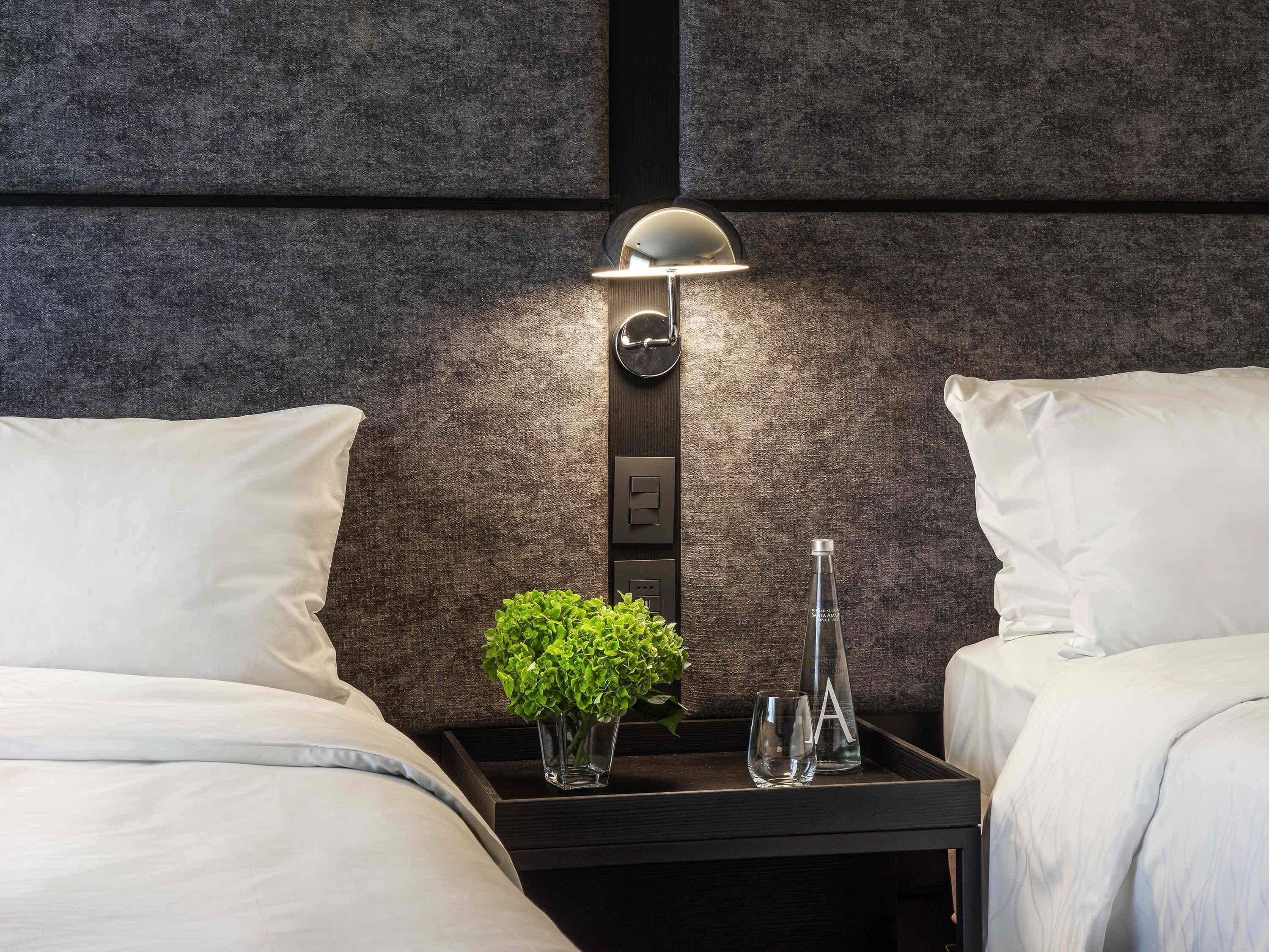 A hotel room with a bed, a nightstand with a lamp, a vase with a plant, and a bottle.