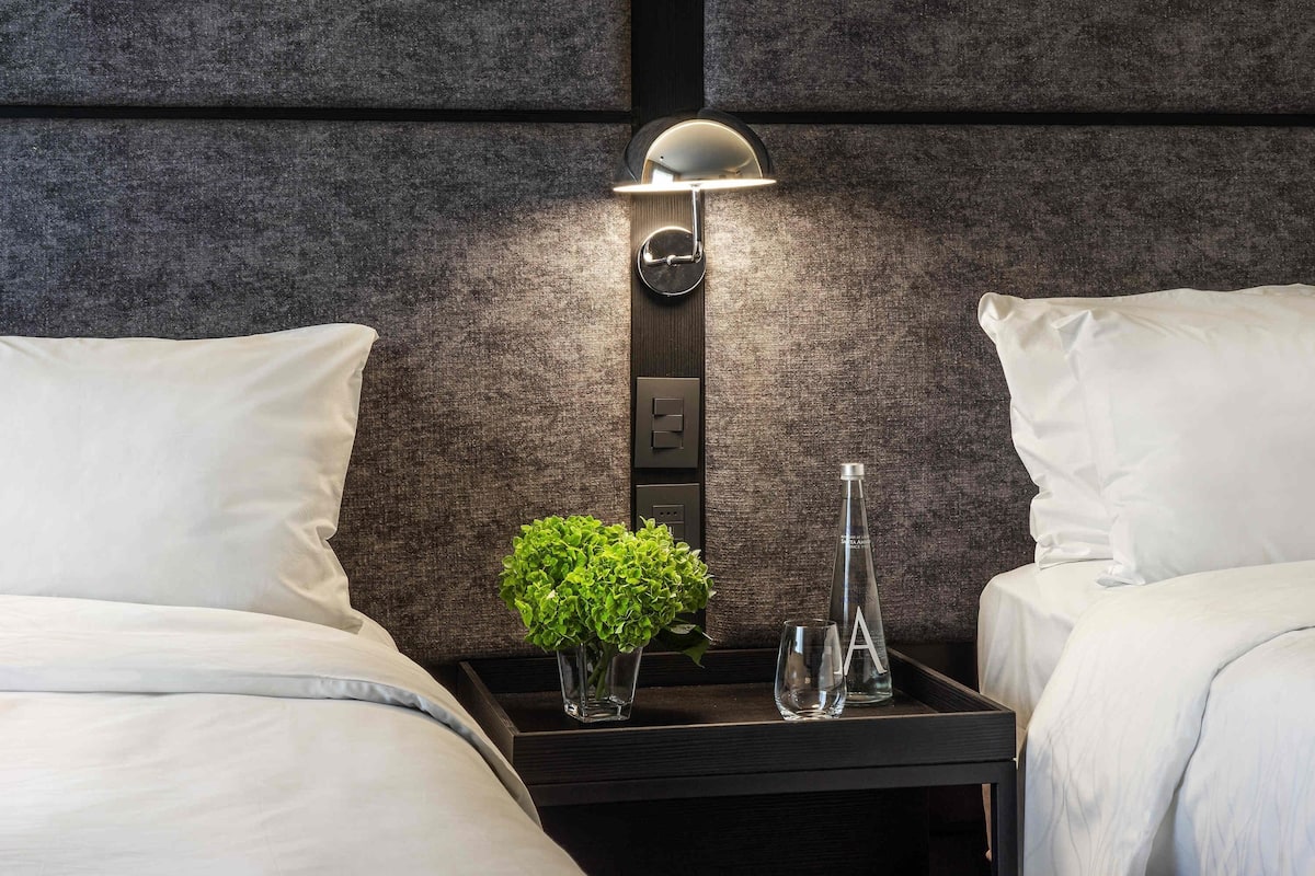 A hotel room with a bed, a nightstand with a lamp, a vase with a plant, and a bottle.