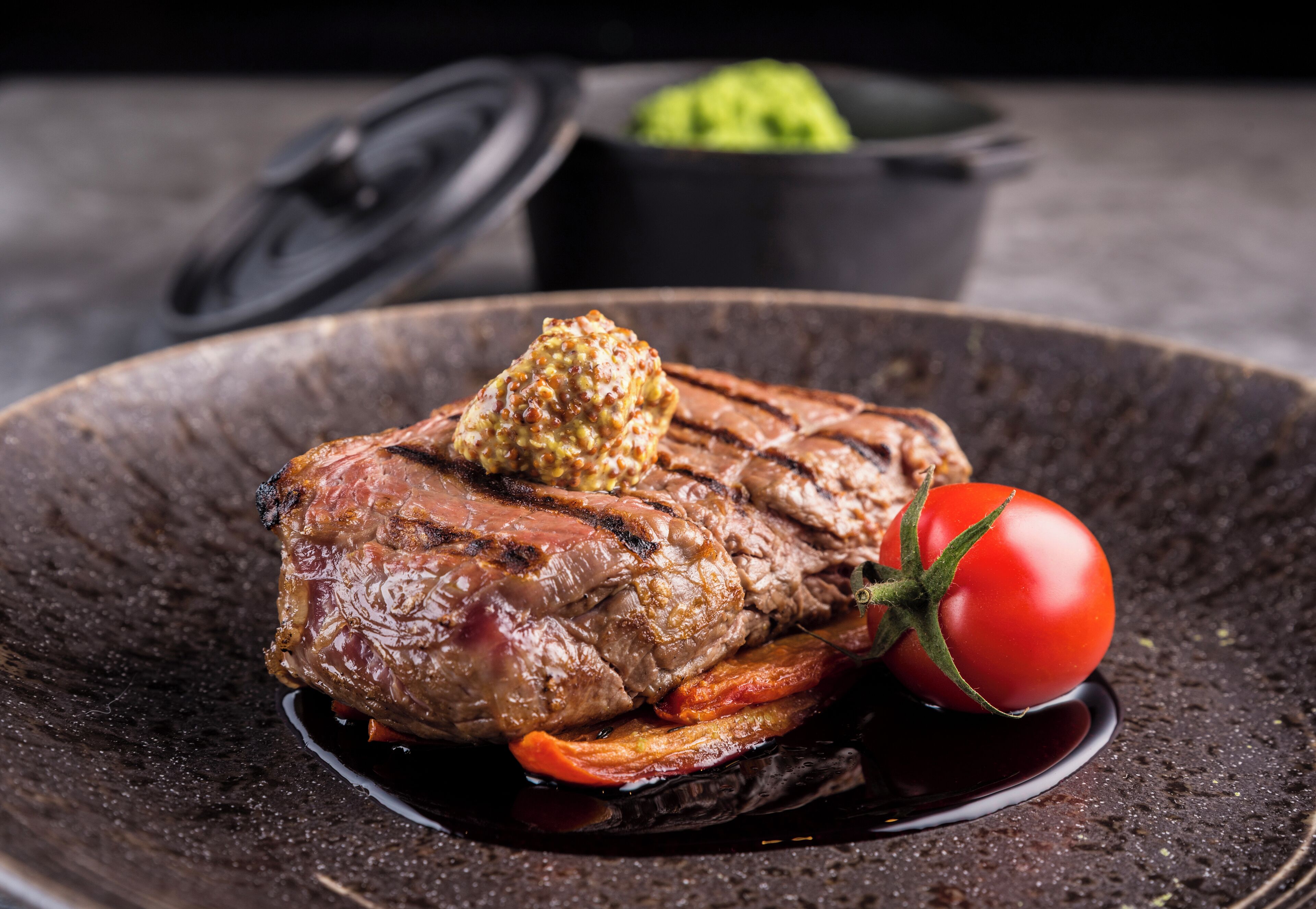 Un bistec con un poco de mantequilla encima, acompañado de un tomate cherry y verduras a la parrilla en un plato oscuro.