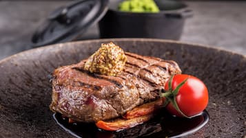Un bistec con un poco de mantequilla encima, acompañado de un tomate cherry y verduras a la parrilla en un plato oscuro.
