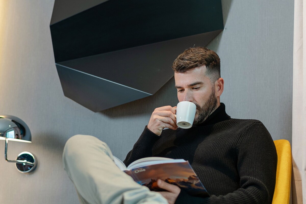 Un hombre sentado en una silla amarilla leyendo un libro, con un diseño moderno de pared geométrica al fondo.