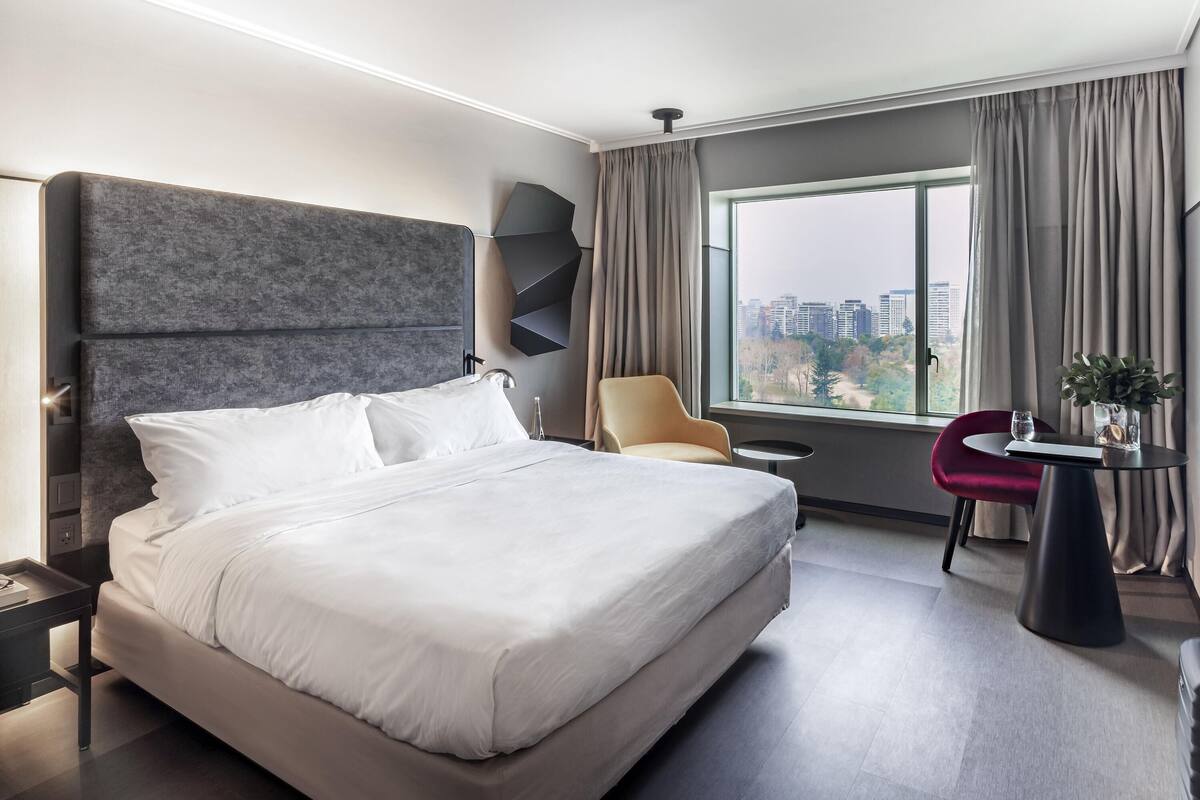 Une chambre d’hôtel moderne avec un grand lit, une chaise, une petite table et une vue sur le paysage urbain par la fenêtre.