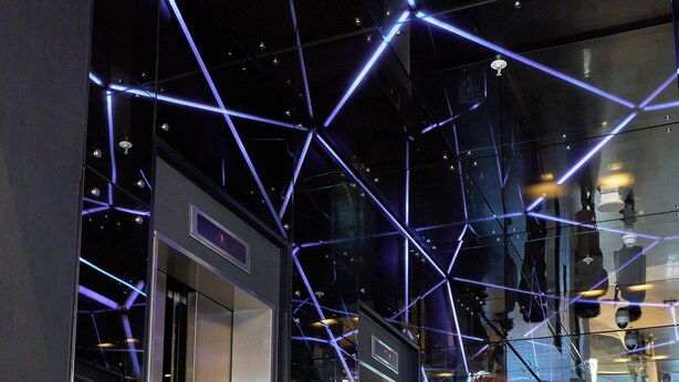 Een interieur met een modern design, met een glazen lift en een geometrisch plafondpatroon.