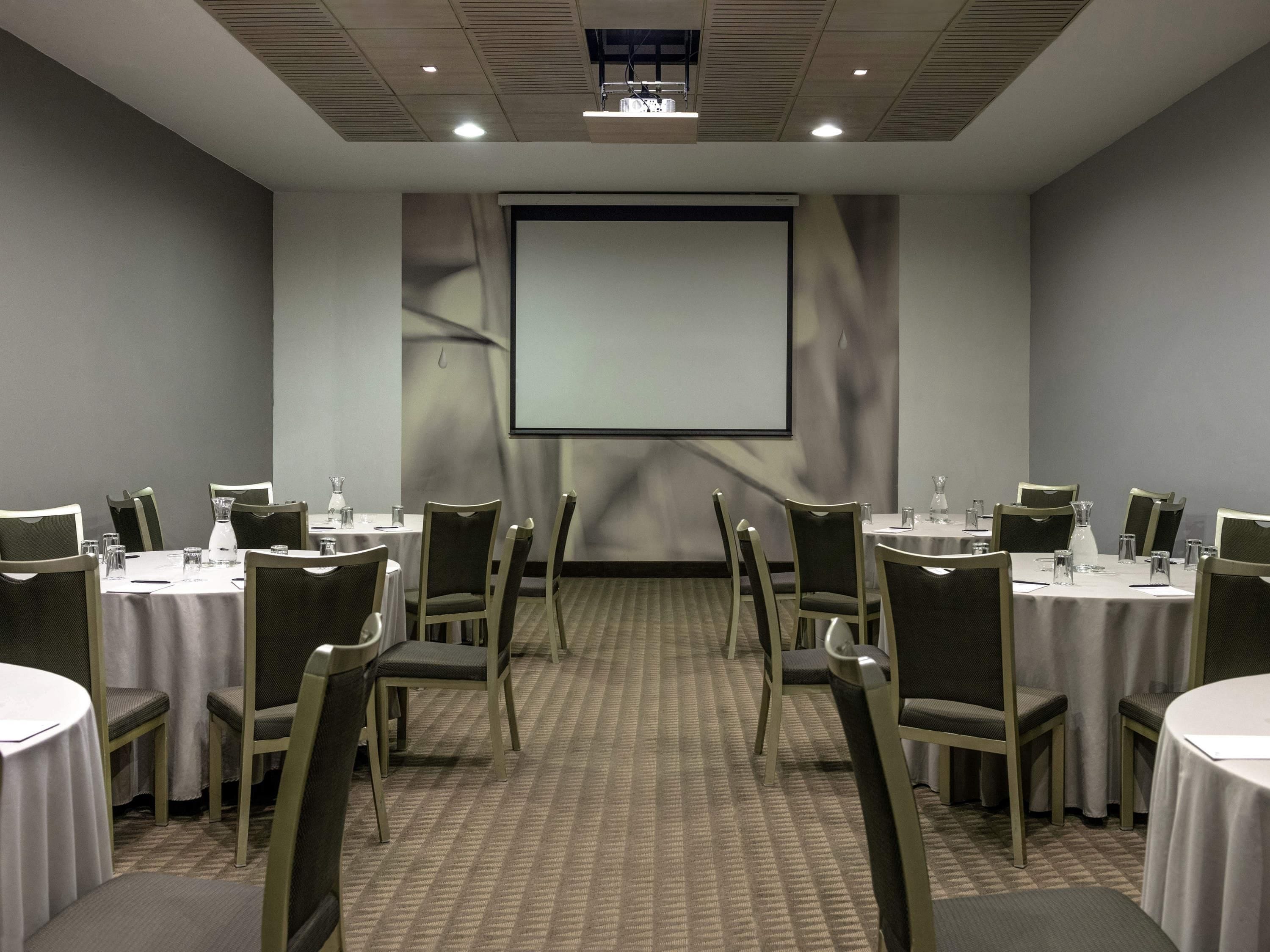Un salón de conferencias con mesas, sillas, una pantalla de proyector y piso alfombrado.