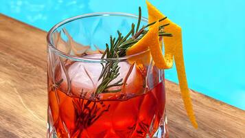A glass with a drink garnished with rosemary and an orange peel, placed on a wooden surface near a pool.