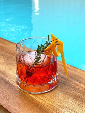 A glass with a drink garnished with rosemary and an orange peel, placed on a wooden surface near a pool. - Pullman Santiago Vitacura (Santiago)