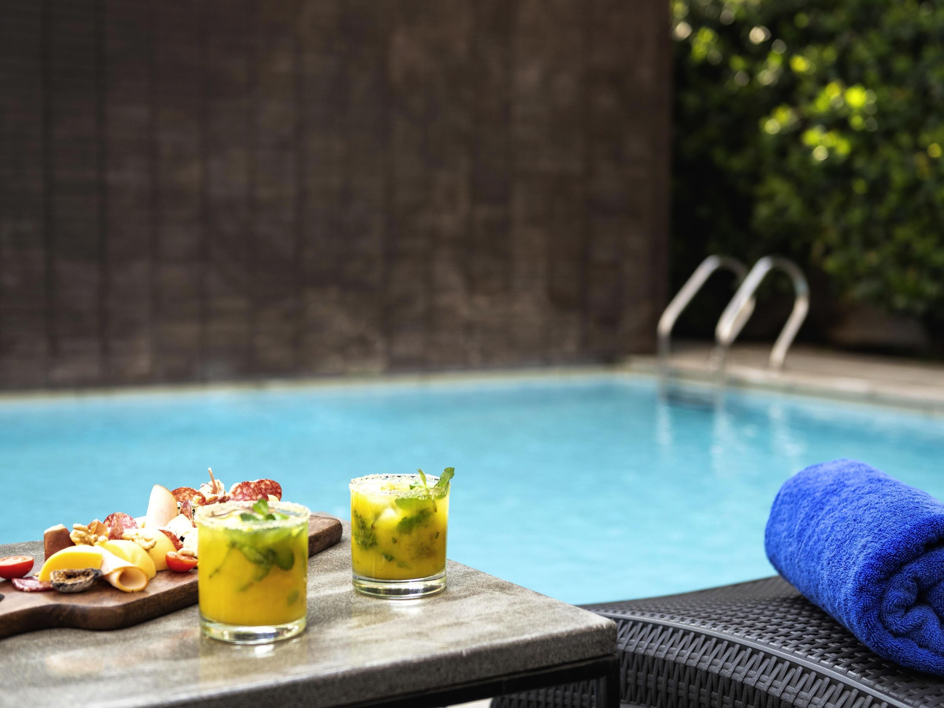 A poolside setting with a dining table featuring a charcuterie board and two drinks, a blue towel, and a pool in the background.