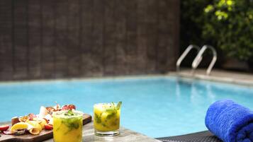 A poolside setting with a dining table featuring a charcuterie board and two drinks, a blue towel, and a pool in the background.