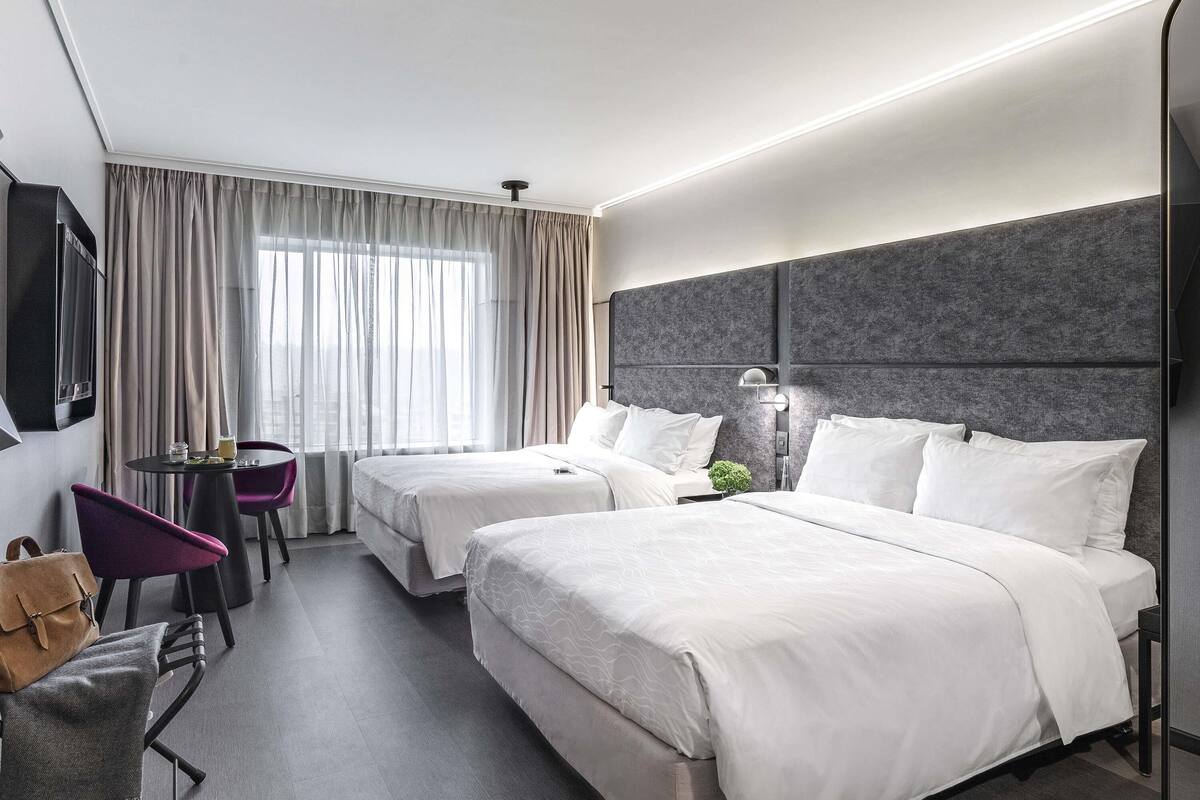 Une chambre d’hôtel avec deux lits, un téléviseur fixé au mur, une petite table avec des chaises et une vue sur la ville grâce à de grandes fenêtres.