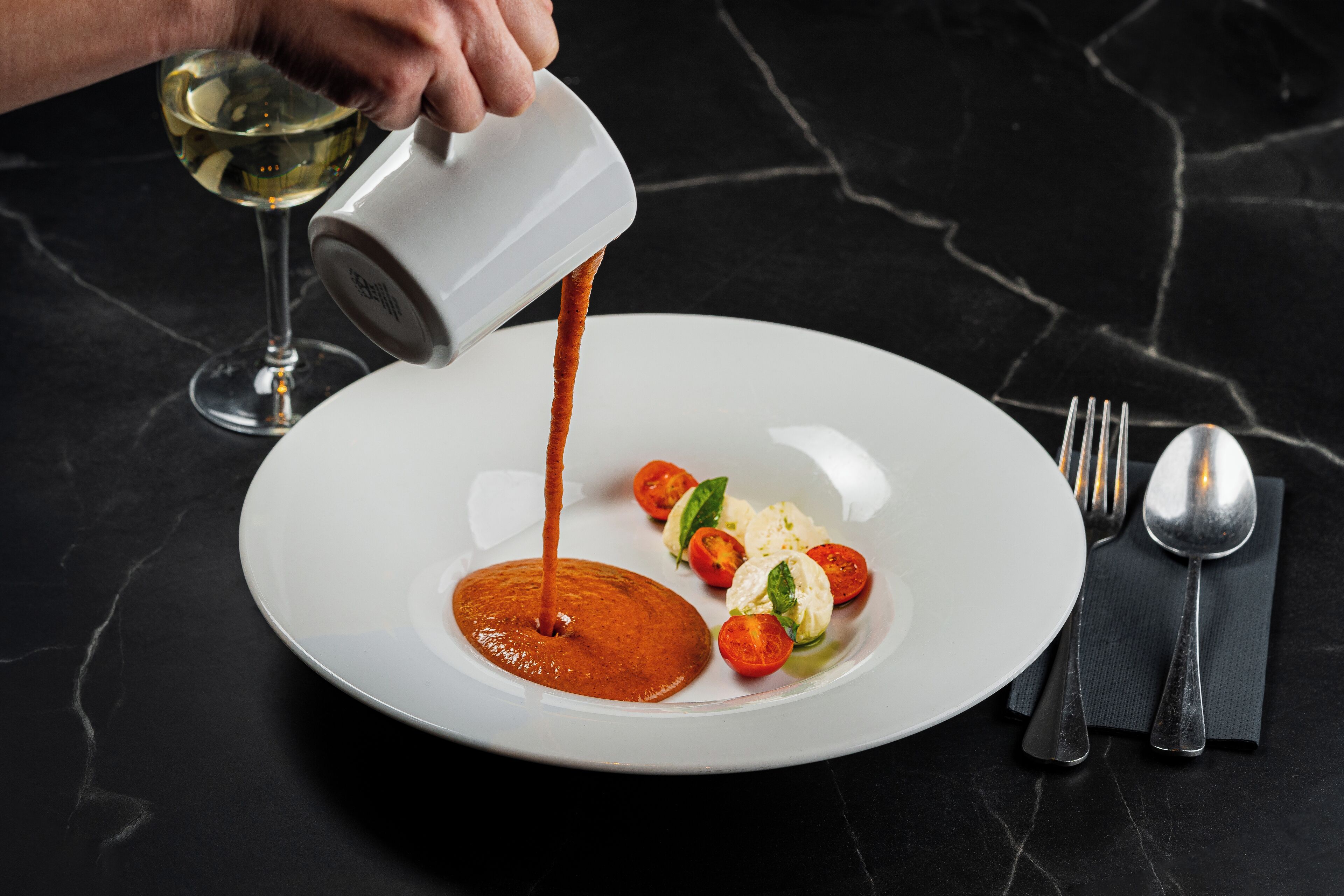 Um prato branco com molho vermelho sendo servido, acompanhado de tomates cereja e folhas de manjericão.
