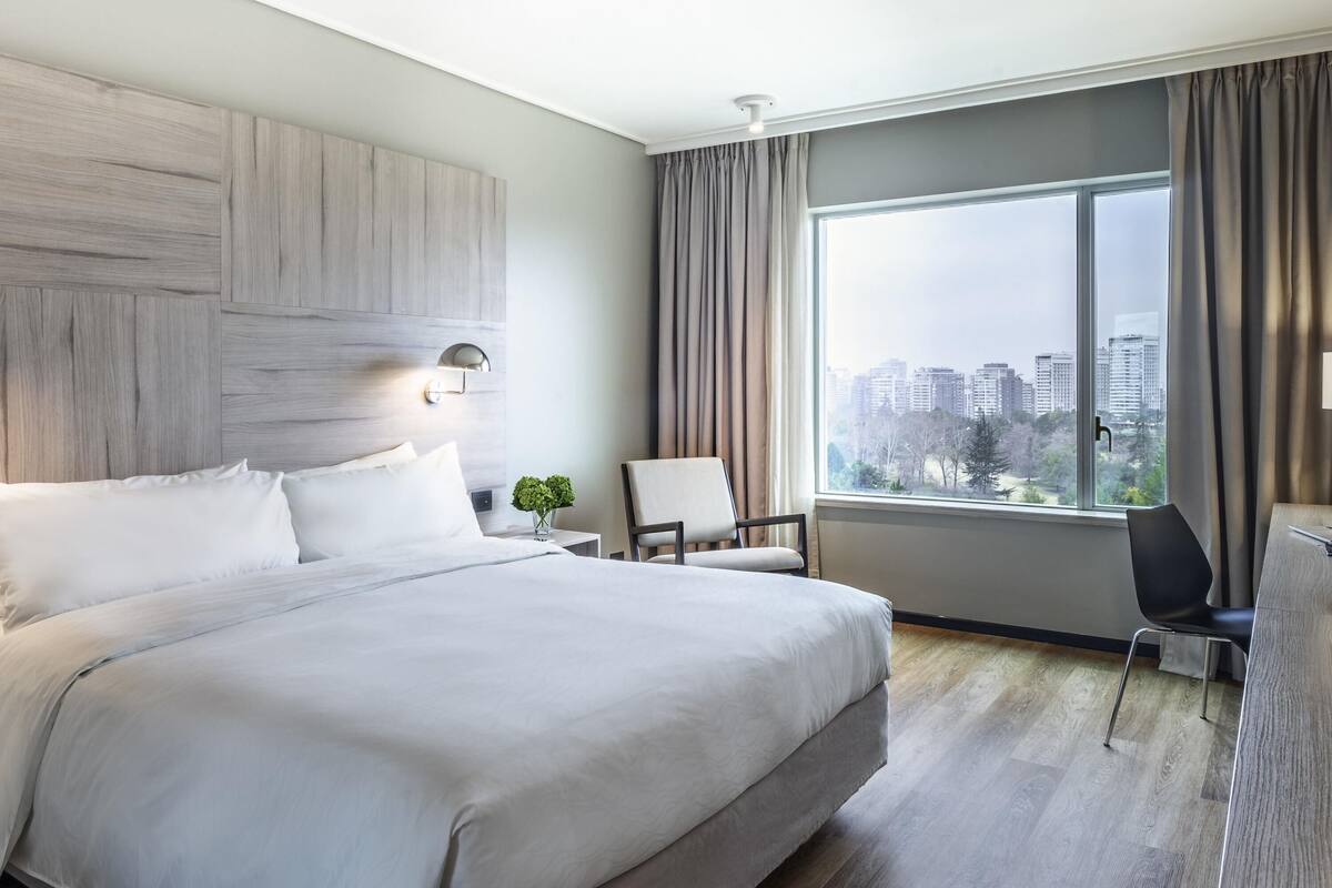 Une chambre d’hôtel moderne, dotée d’un grand lit, d’un bureau, d’une chaise et offrant une vue sur le paysage urbain.