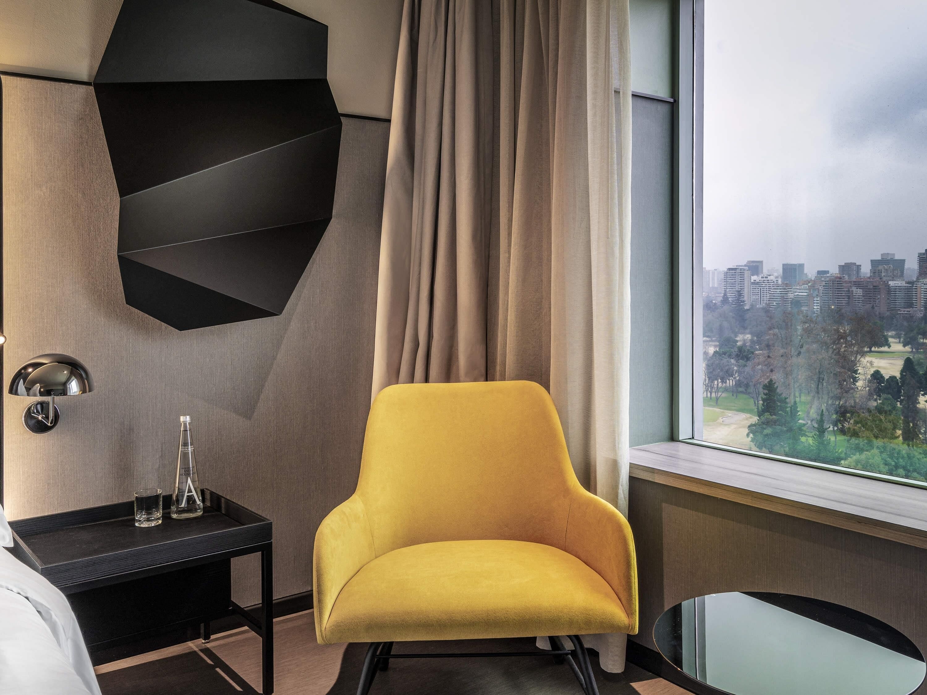 Une chambre d’hôtel moderne avec un fauteuil jaune, une table d’appoint noire sur laquelle se trouve un verre, et une vue sur la ville par la fenêtre.