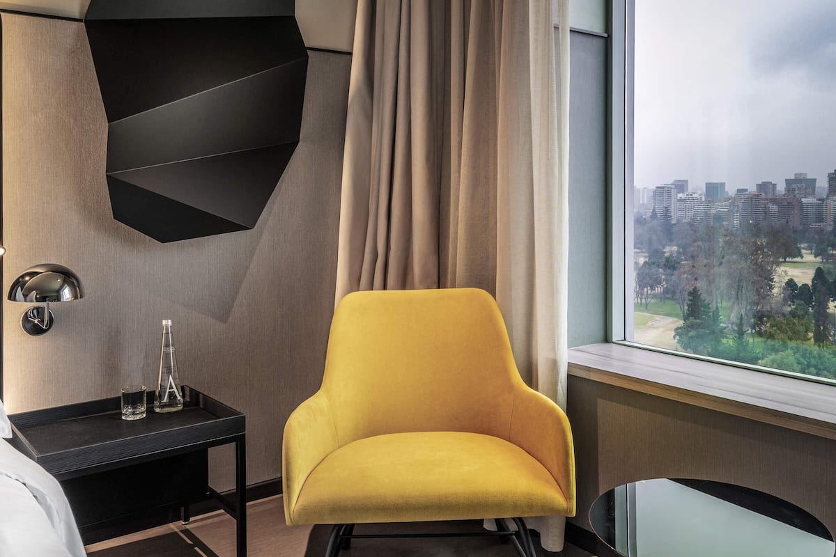 A modern hotel room with a yellow armchair, a black side table with a glass, and a city view through the window.