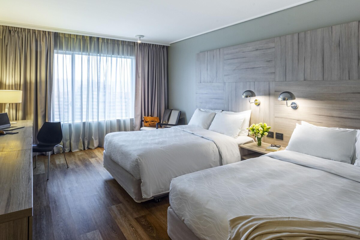 Habitación de hotel con dos camas, un escritorio, una silla, una lámpara y vistas al exterior.