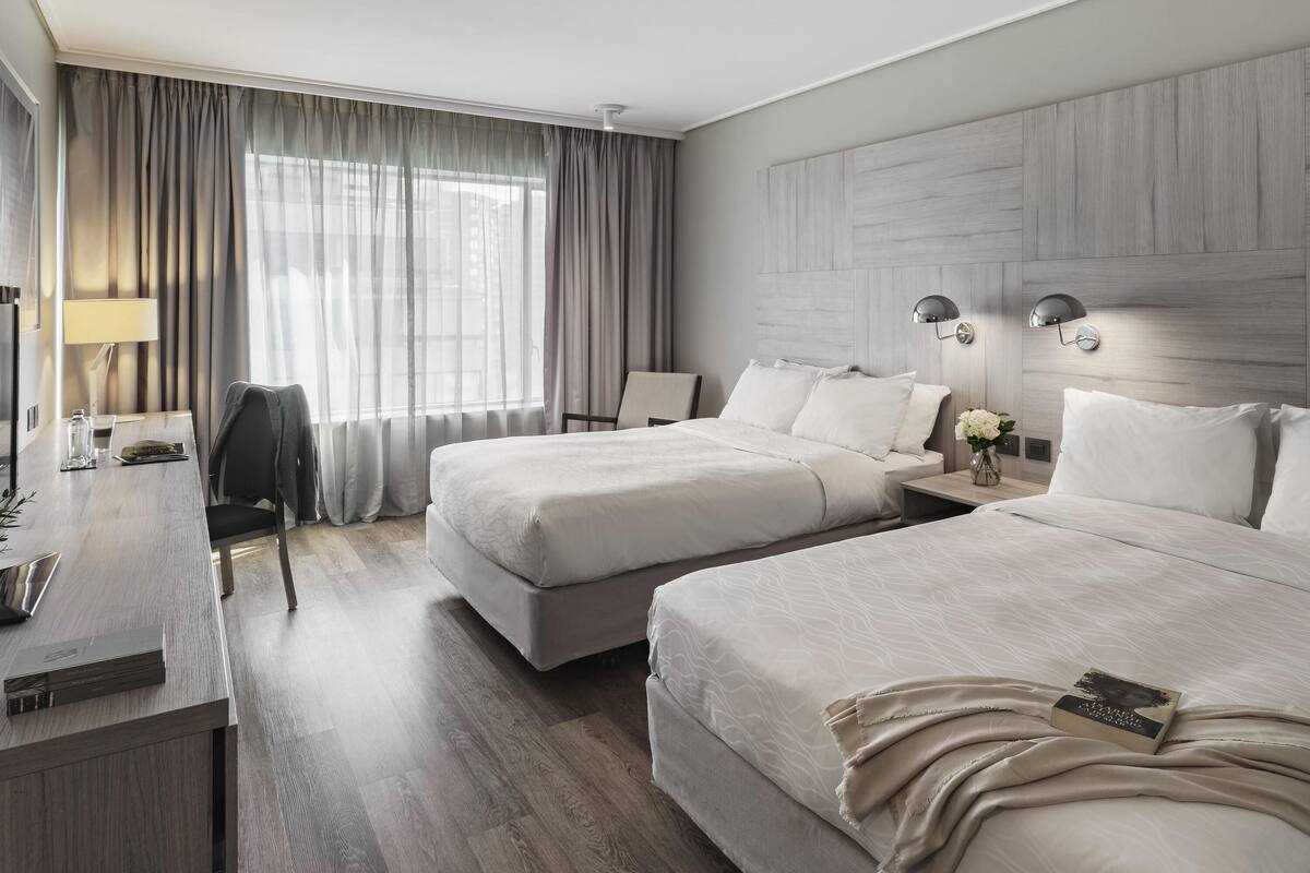 Une chambre d’hôtel avec deux lits, un bureau, une chaise, une lampe et une fenêtre avec des rideaux.