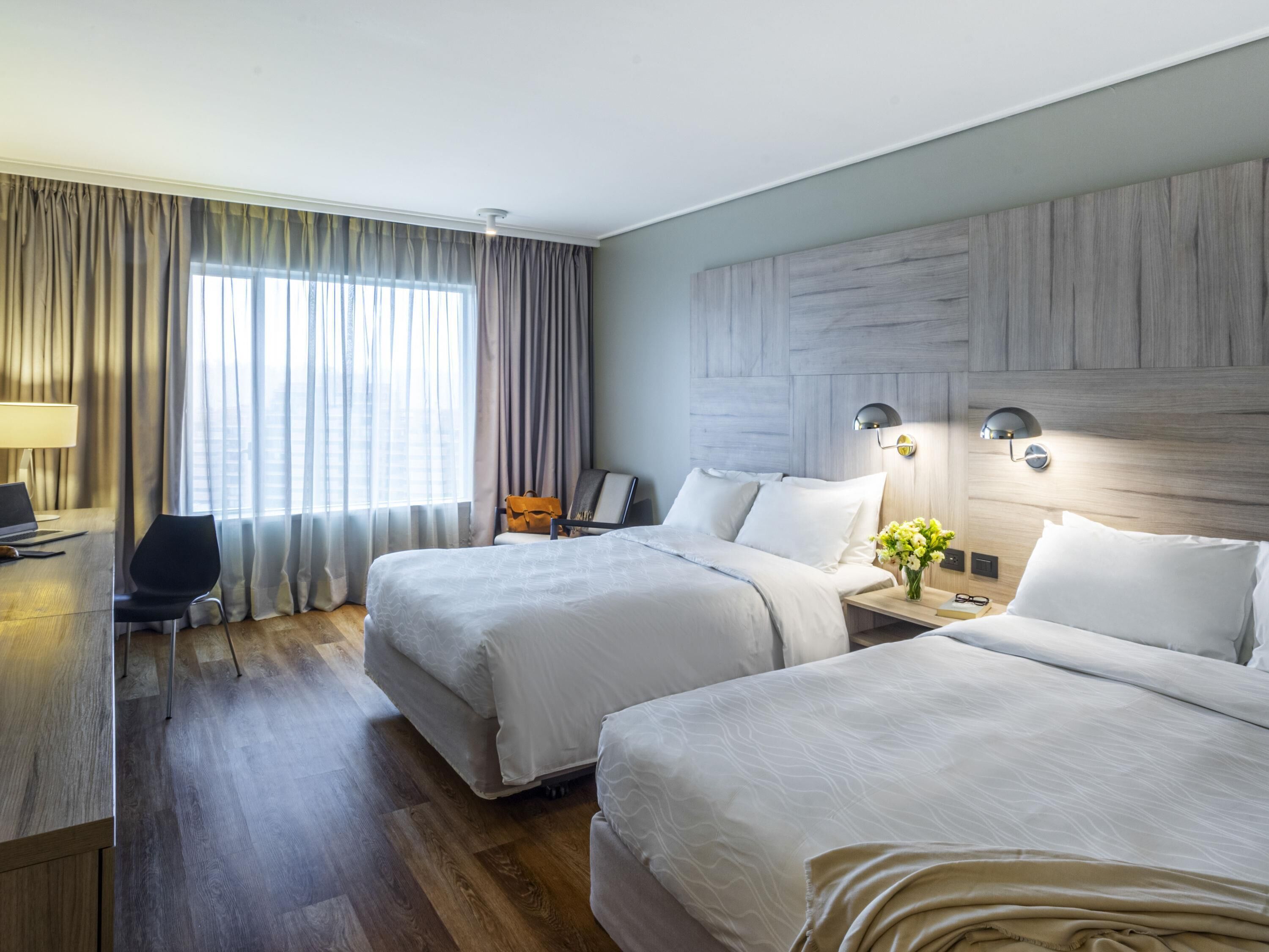 Une chambre d’hôtel avec deux lits, un bureau, une chaise, une lampe et une vue sur l’extérieur.
